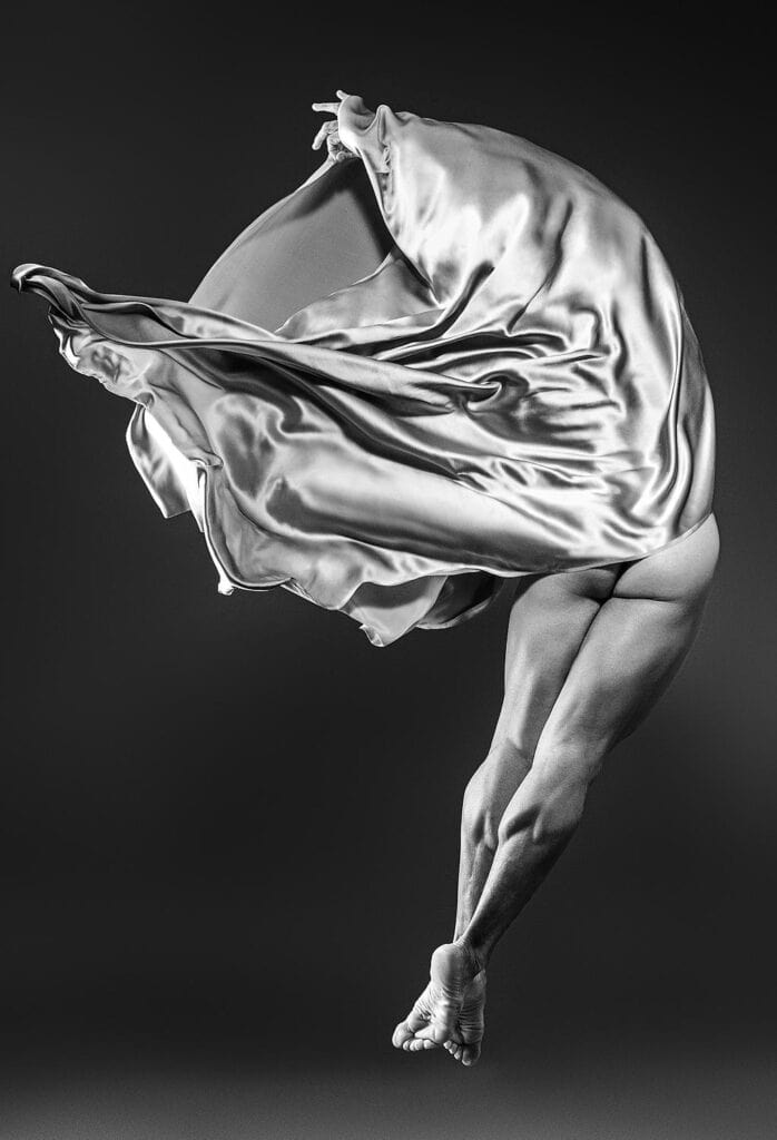 A partially obscured figure bends beneath billowing satin fabric, with crossed legs and bare feet against a dark background.
