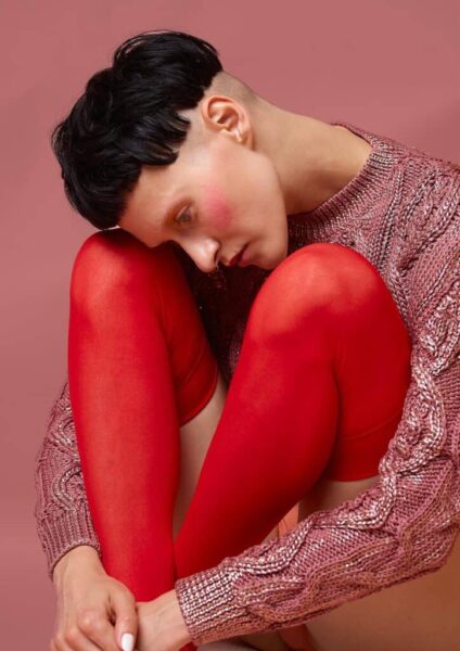 Cropped portrait of a seated person in red tights and a rose-toned knit sweater against a muted pink background.