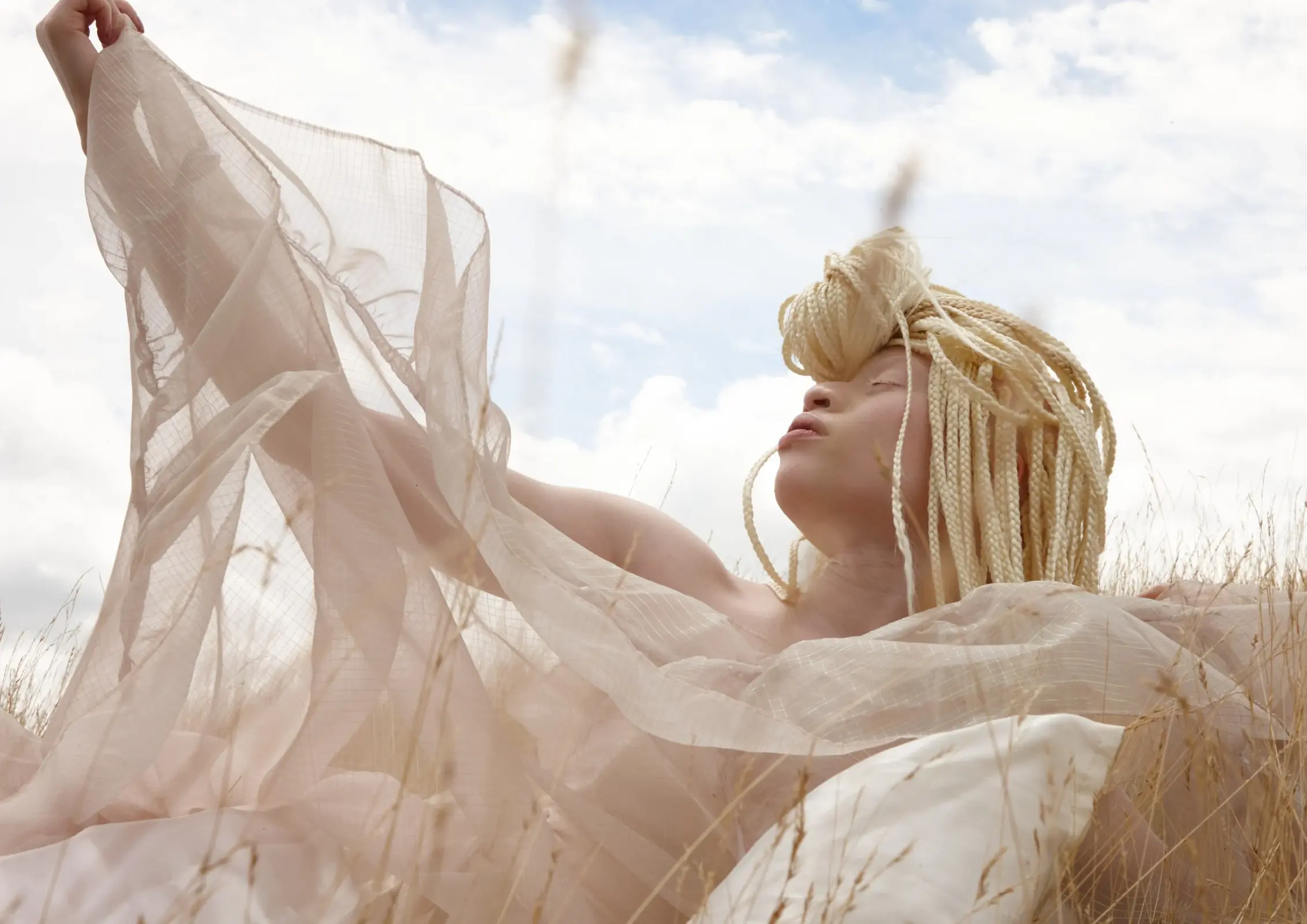 Blonde model reclining outdoors while lifting translucent pale fabric against a bright sky.
