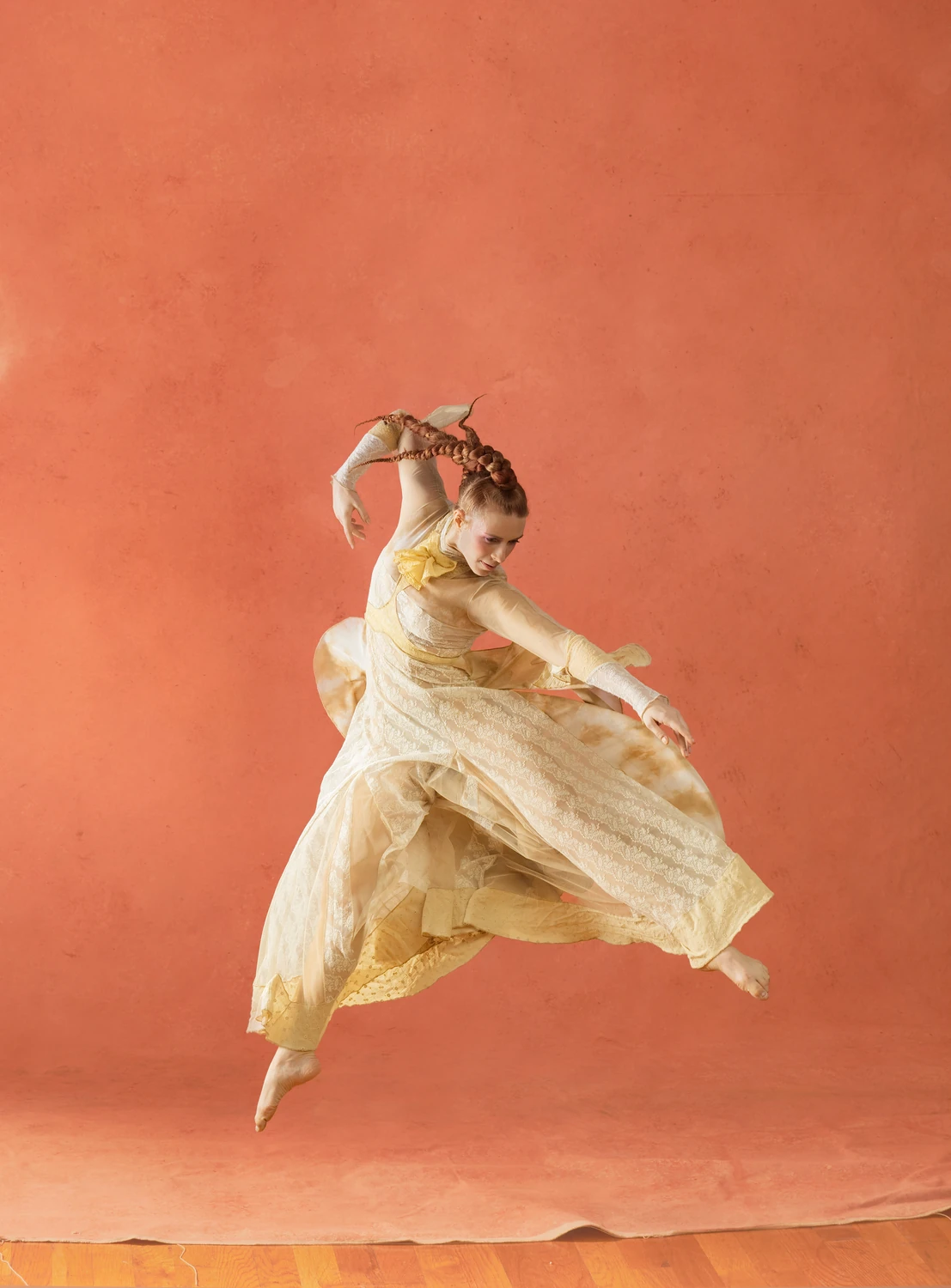 Performer airborne in a leap as layered cream garments flare against the terracotta background.