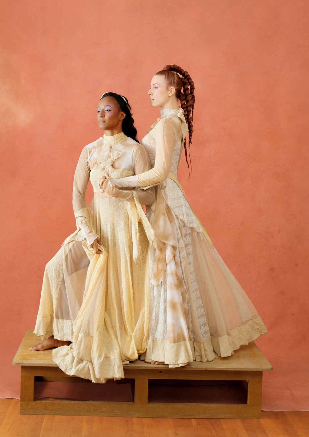 Two performers in layered cream garments standing together on a low wooden platform against terracotta.