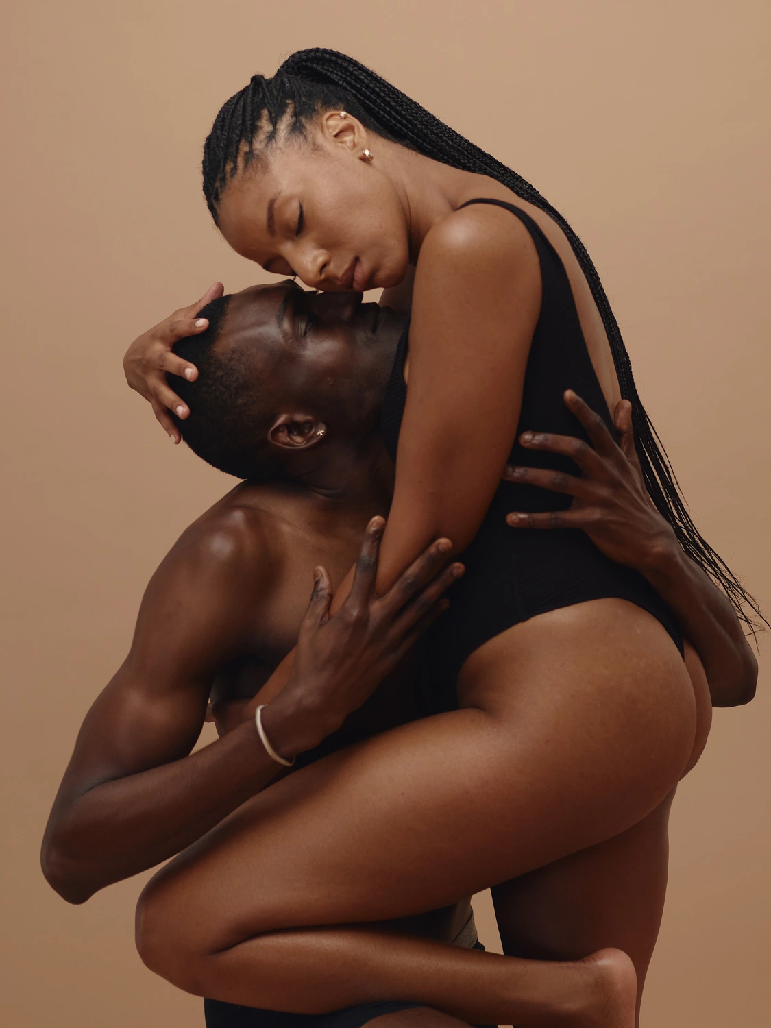 Two people in a sculptural seated embrace against a warm neutral studio background.