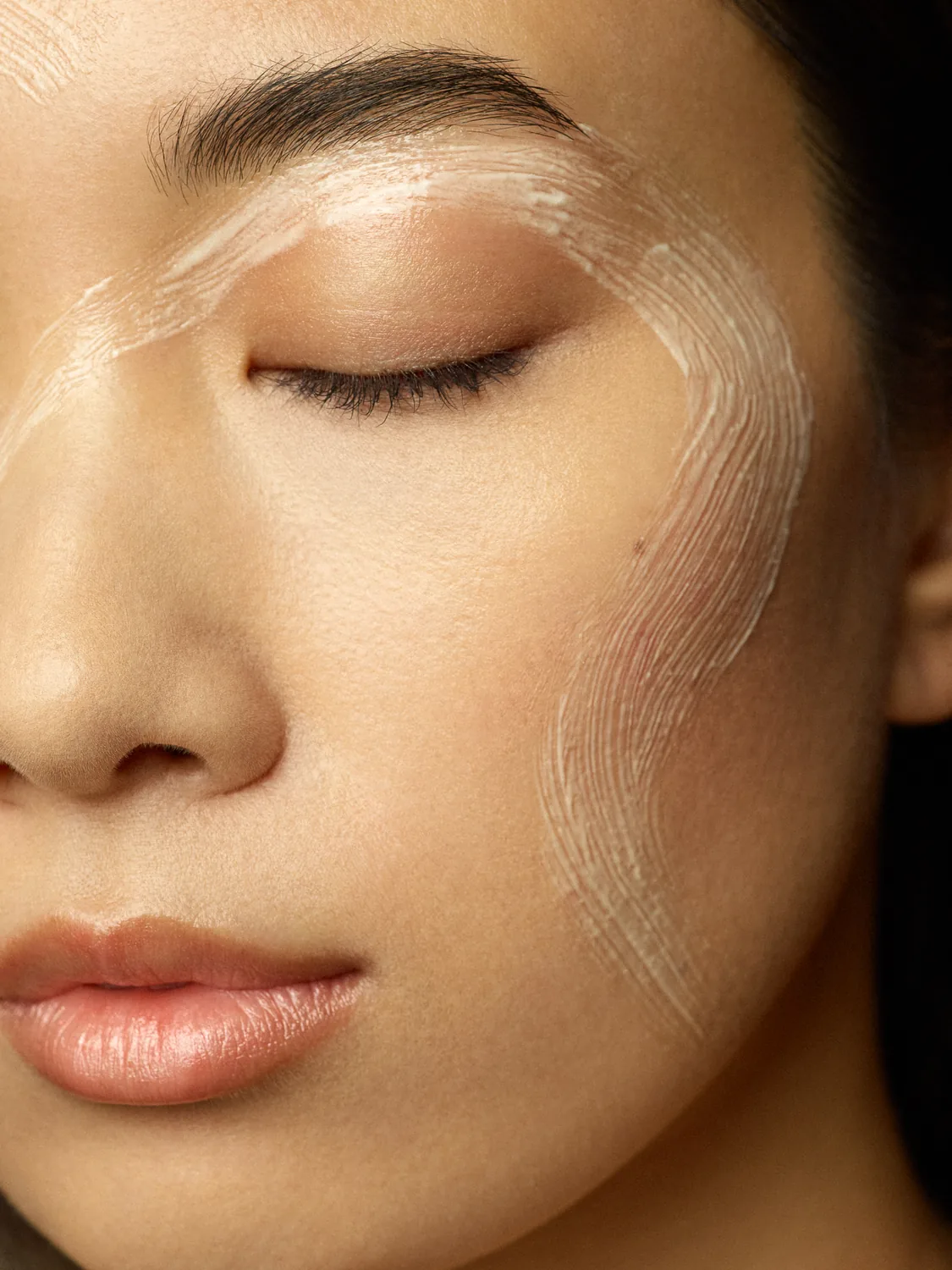 Close beauty crop with eyes closed and a translucent skincare mask brushed across the cheek and eye area.