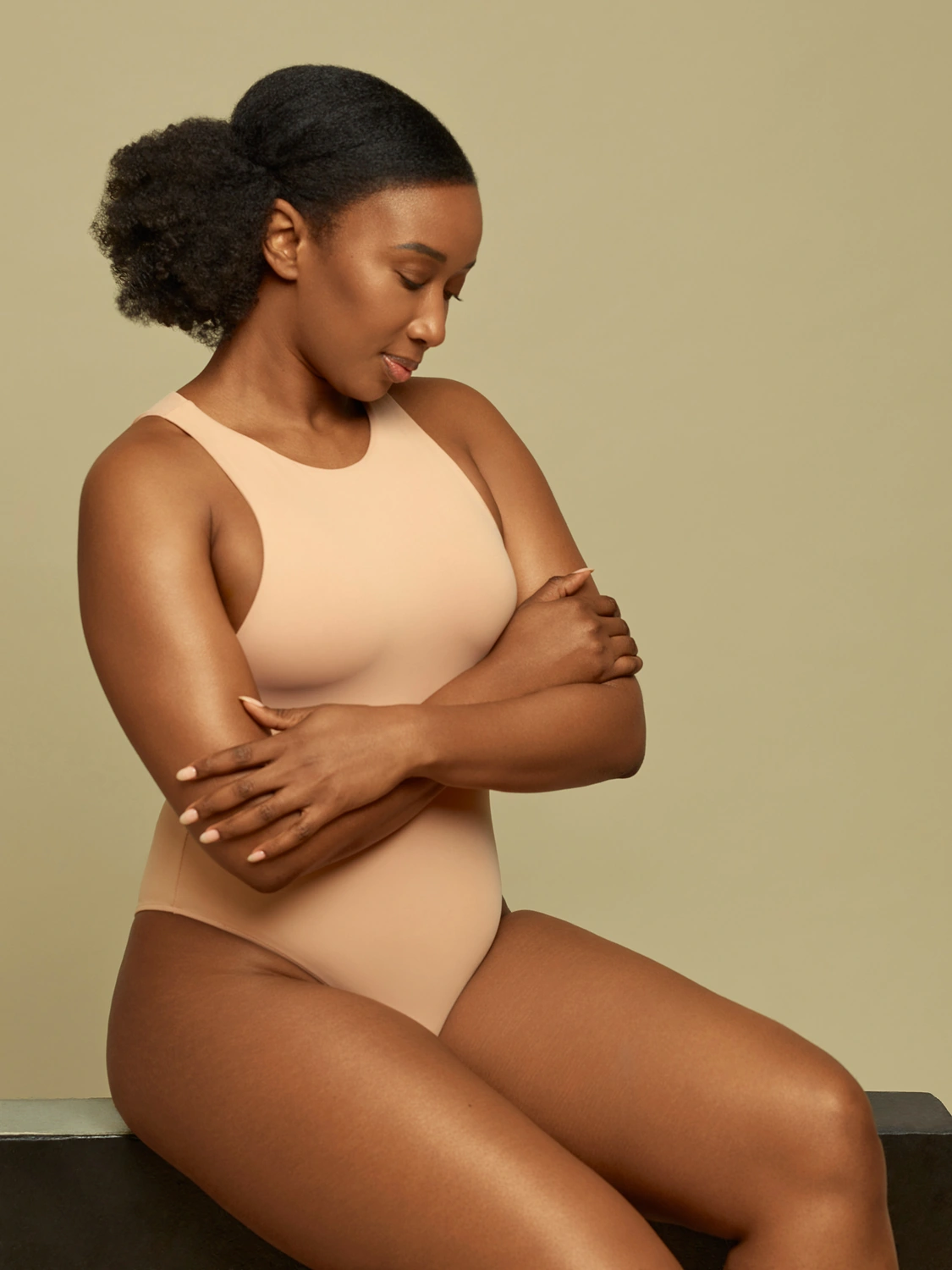 Seated beauty portrait of a dark-skinned woman in a neutral bodysuit against a warm beige background.