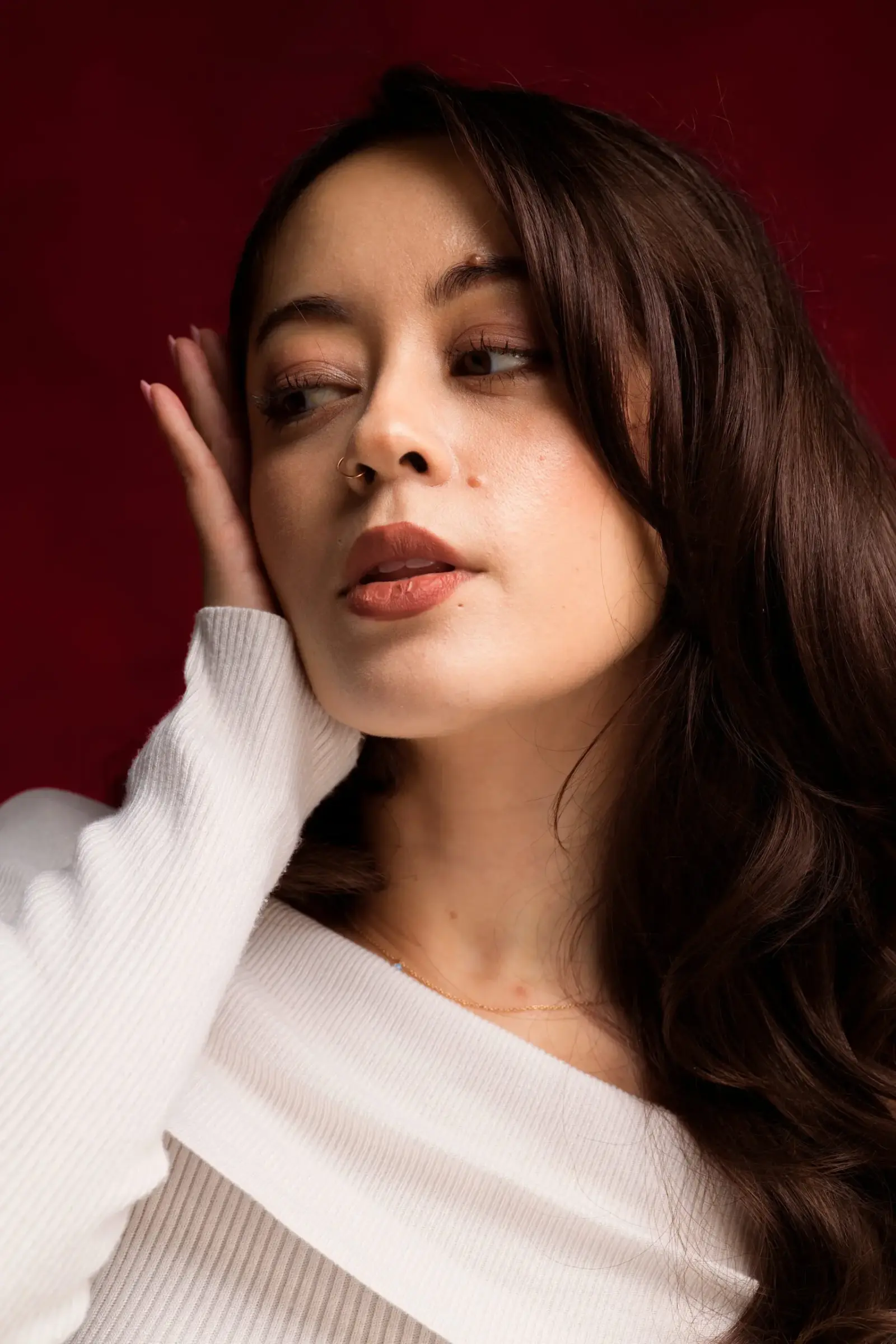 Close-up portrait of a person in a white ribbed sweater, resting one hand beside the face against a deep red backdrop.