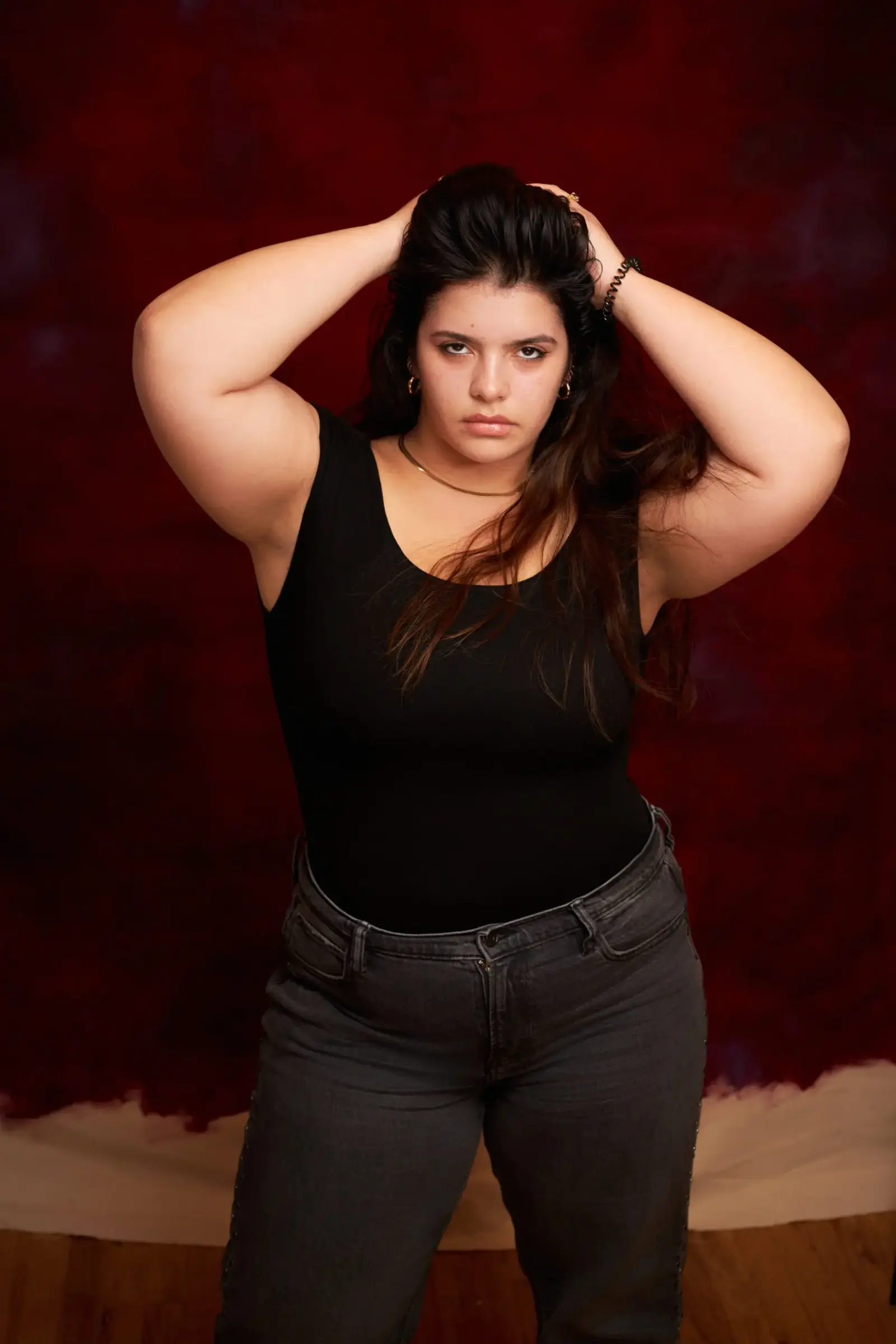 Person standing against a red-painted backdrop with both hands raised to their hair, wearing a black top and dark jeans.