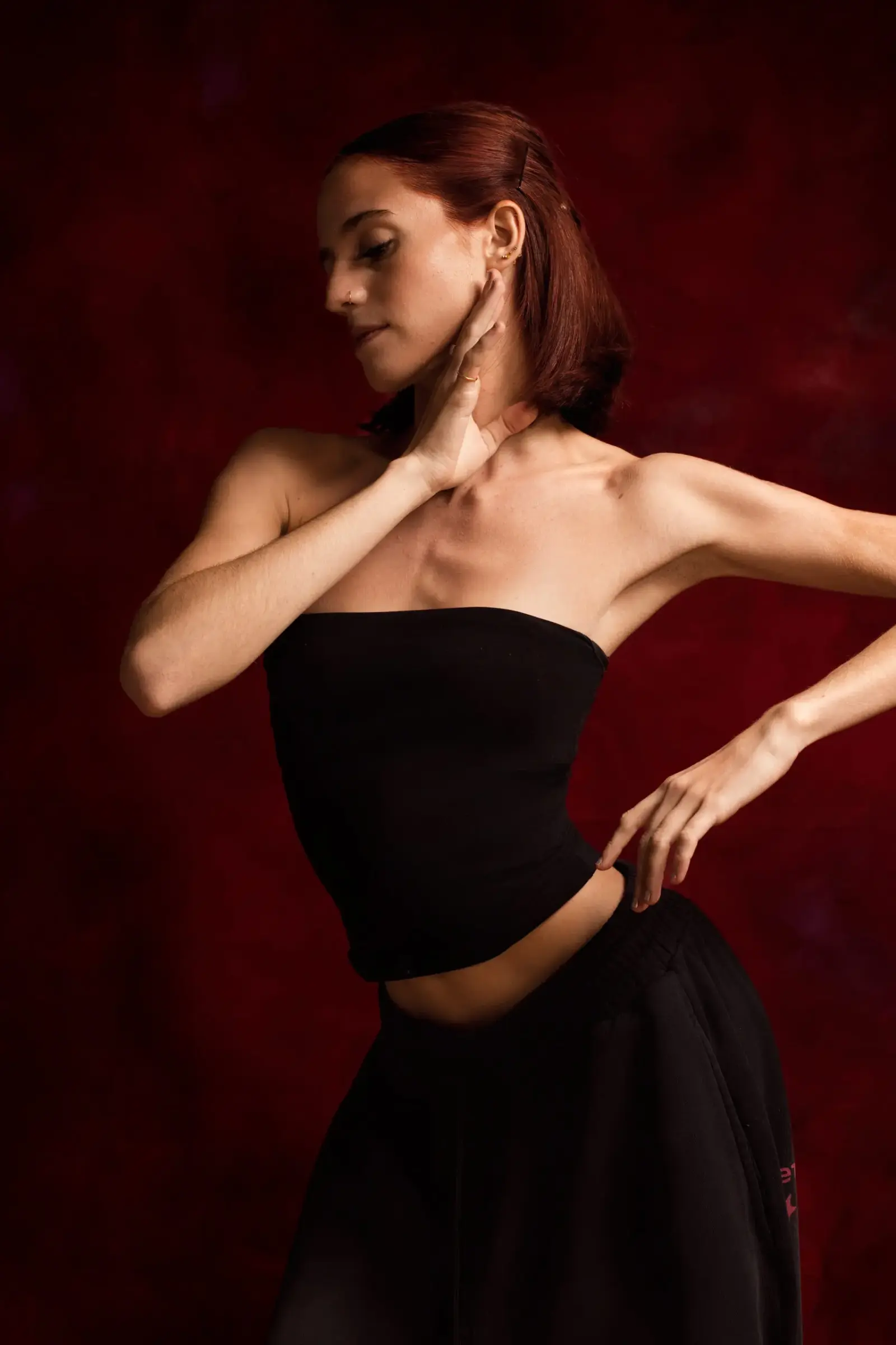 Portrait of a person in a black strapless top and trousers, posing with one hand at the neck against a red backdrop.